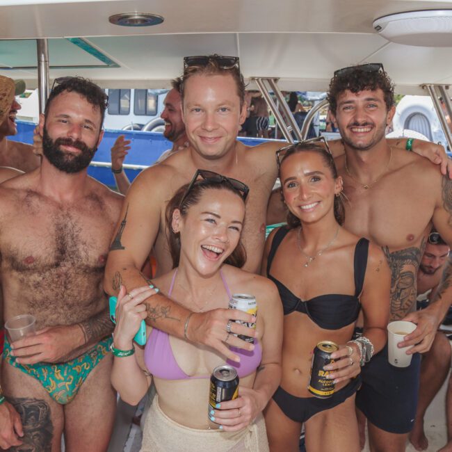 A group of smiling young adults in swimsuits pose together on a boat, holding drinks. They appear to be enjoying a party, with other people and boats visible in the background.