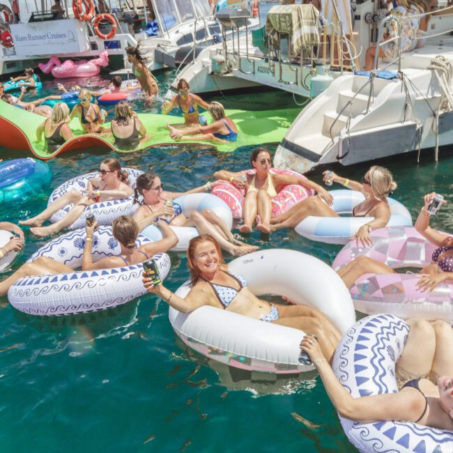 A group of people relax on colorful inflatable pool floats in the sea near anchored boats, enjoying a sunny day. Some are smiling at the camera while others chat and sunbathe in the water.