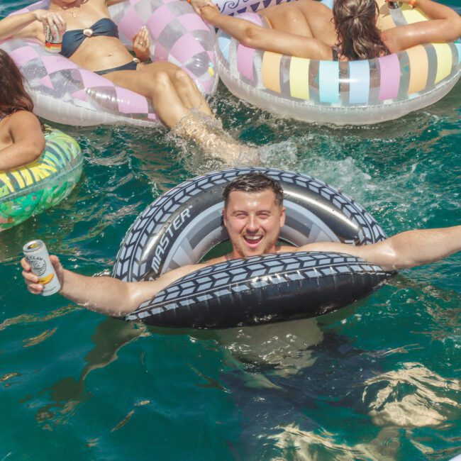 A man in a tire-shaped pool float smiles and gives two thumbs up while holding a can, surrounded by other people relaxing on colorful inflatables in bright blue water.