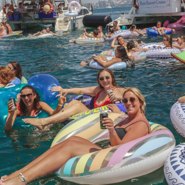 A group of people relax on colorful inflatable tubes in the water near boats, smiling and holding drinks, enjoying a sunny day at a lively floating party. The atmosphere is festive and social.