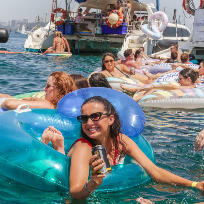 A group of people relax on colorful pool floats in the sea near anchored yachts. A smiling woman in sunglasses holds a drink, enjoying the sunny day and lively atmosphere with others around her.