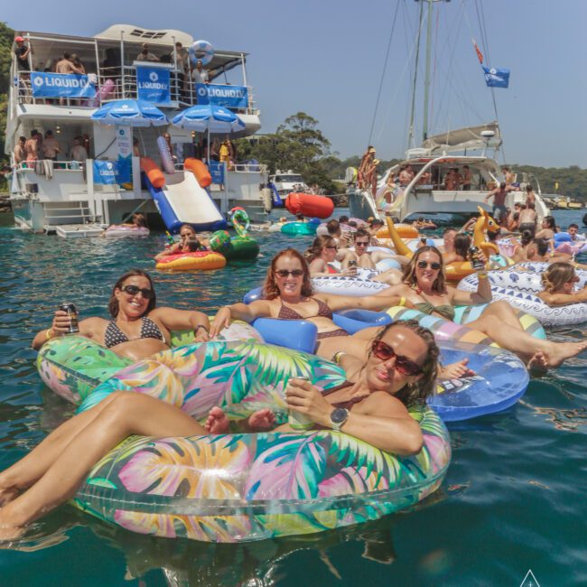 A group of people relax on colorful inflatables in the water near anchored yachts during a sunny day, enjoying drinks and socializing at a lively boat party.