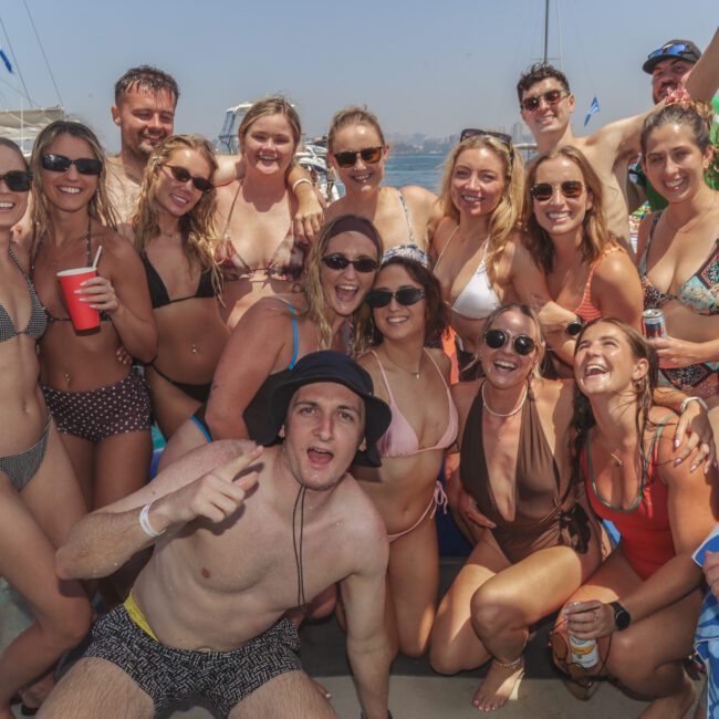 A group of people in swimsuits pose together on a boat, smiling and enjoying a sunny day on the water with sailboats and a city skyline in the background.