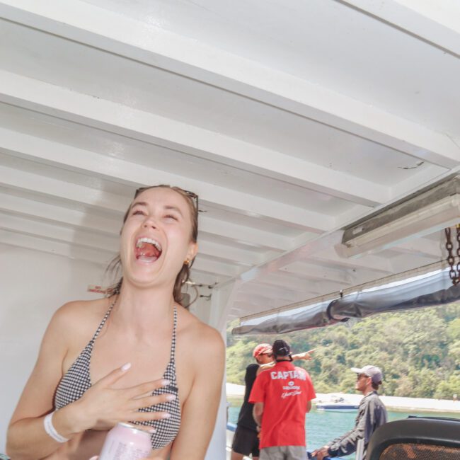 A woman in a checkered bikini laughs while holding a drink can on a boat, with people in red shirts and hats visible in the background near the railing and a scenic shoreline beyond.