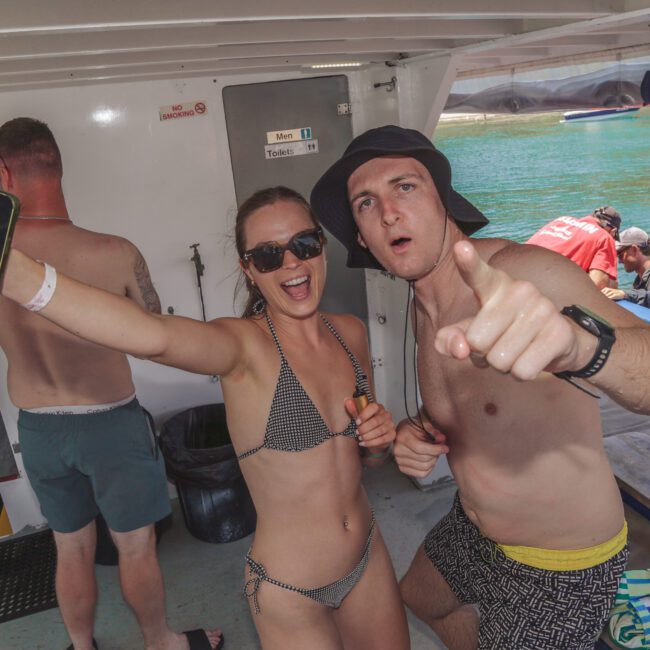Two people in swimsuits, one wearing sunglasses and the other a hat, pose and smile enthusiastically on a boat. Water and more people are visible in the background.