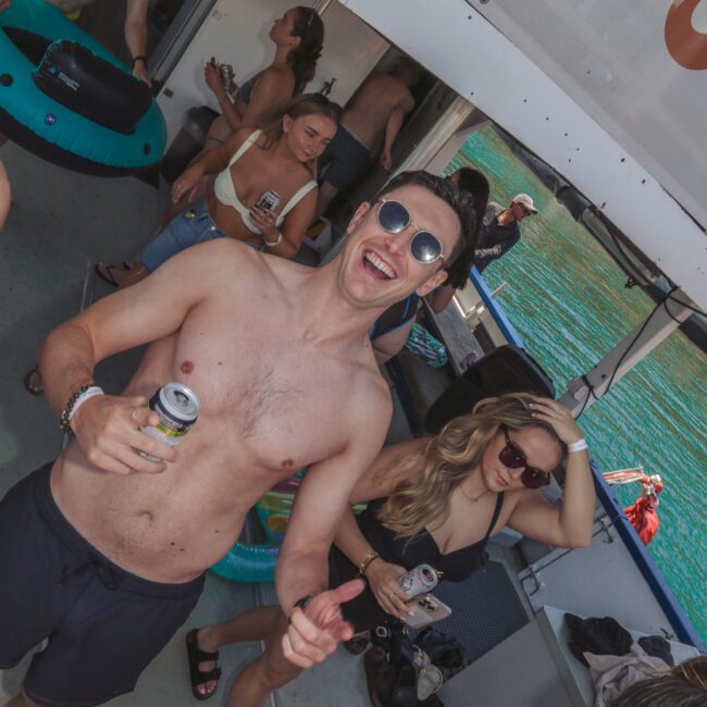 A group of young adults in swimwear enjoy drinks and socialize on a boat, with one man in sunglasses smiling and gesturing to the camera. Sunlight streams through, and water is visible outside the window.
