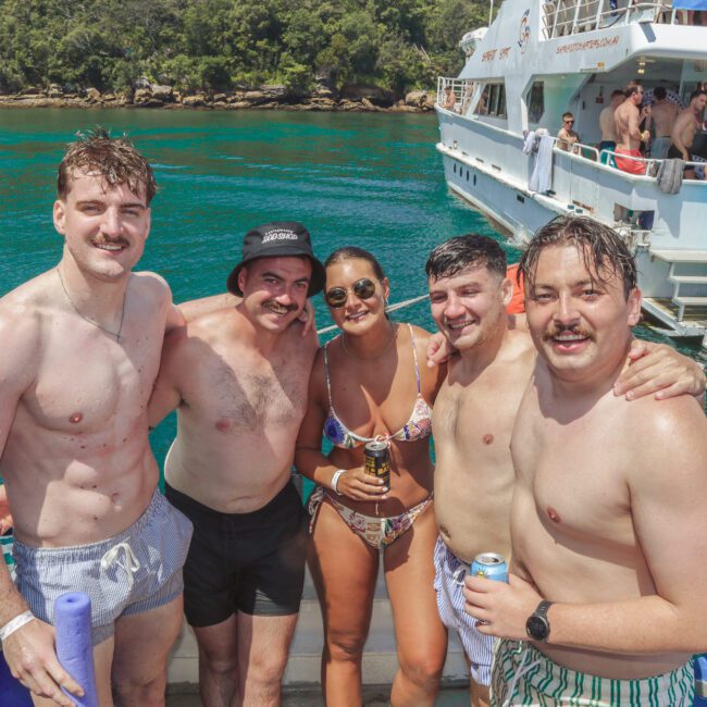 Five people in swimsuits smile for a group photo on a boat, with turquoise water and another yacht in the background. The group appears to be enjoying a sunny day on the water.