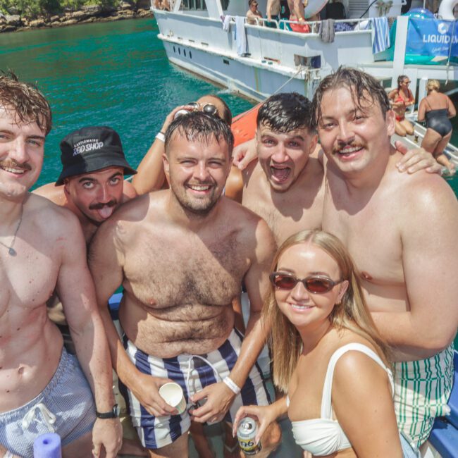A group of five smiling men in swimwear and one woman in sunglasses pose together on a boat under sunny weather, with turquoise water and a white boat in the background.