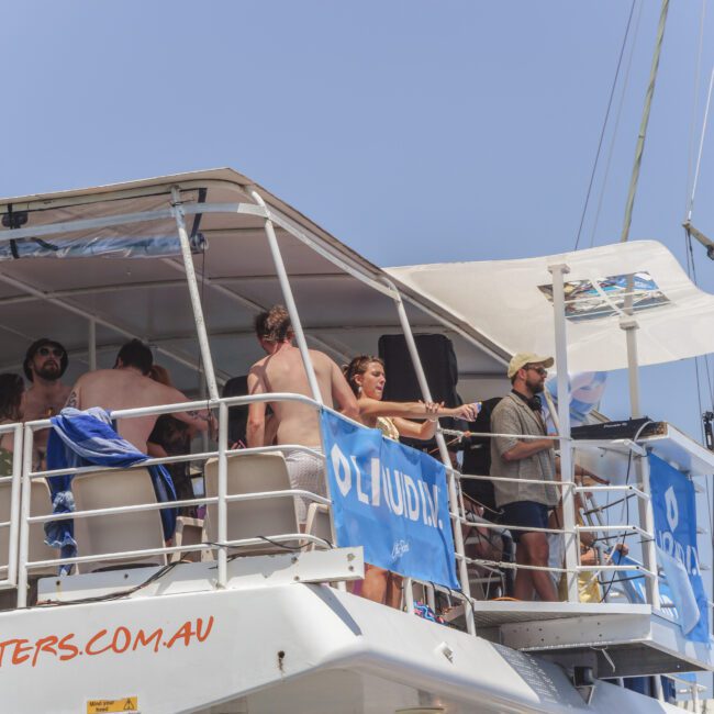 A group of people in swimsuits are standing and socializing on the upper deck of a white boat under clear blue skies. Blue banners and open railings are visible, and some people appear to be enjoying drinks.