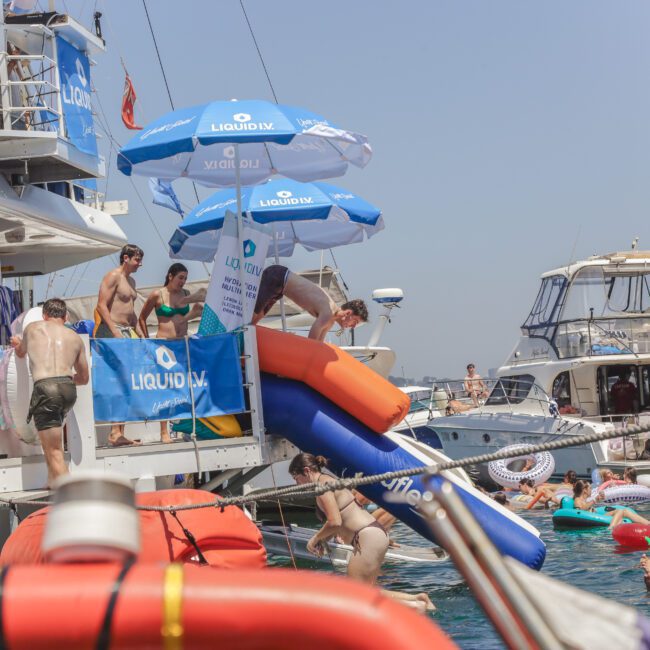 People enjoying a sunny day on boats, swimming, using a waterslide, and relaxing on inflatables in clear blue water. There are branded umbrellas reading "Liquid IV" and several boats docked closely together.