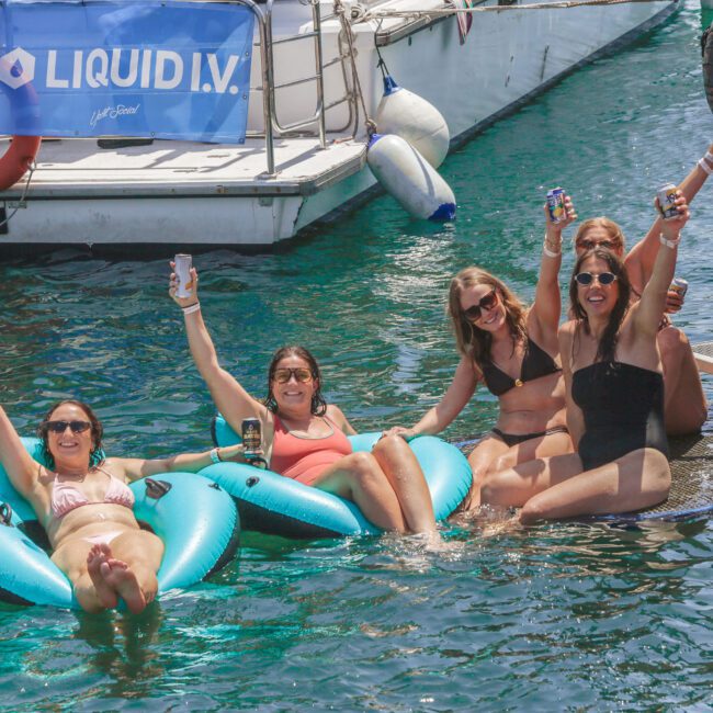 Five women in swimsuits smile and raise drinks while relaxing on floats and a platform in the water near a docked boat. The scene is sunny and festive, with a “LIQUID I.V.” banner visible in the background.