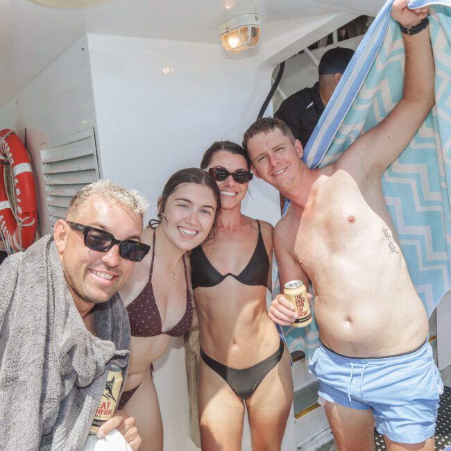 Four smiling adults in swimwear pose on a boat, two holding drinks. One man has a towel draped over his shoulders, another holds up a blue-striped towel. Pool floats and life preservers are visible in the background.