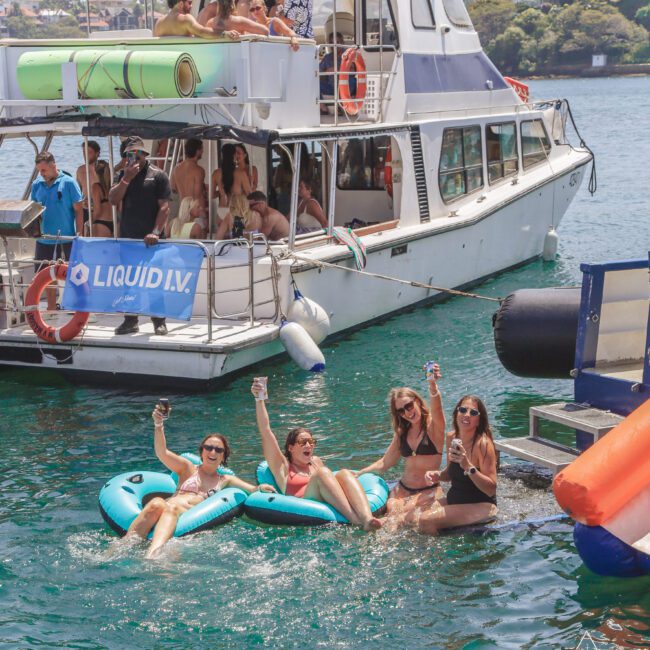 Four women in swimsuits relax on inflatable floats in the water, holding up drinks and smiling. Behind them, people gather and party on a docked boat in sunny weather, with buildings visible in the background.