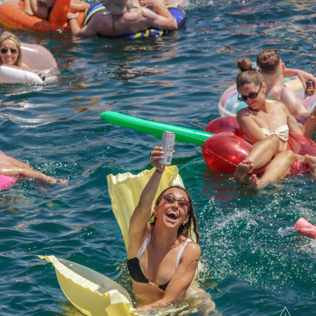 A woman in a black and white bikini raises a can and smiles while sitting on a yellow float in a crowded pool; others on colorful inflatables relax and chat in the background. "Yacht Social Club" logo is visible in the corner.