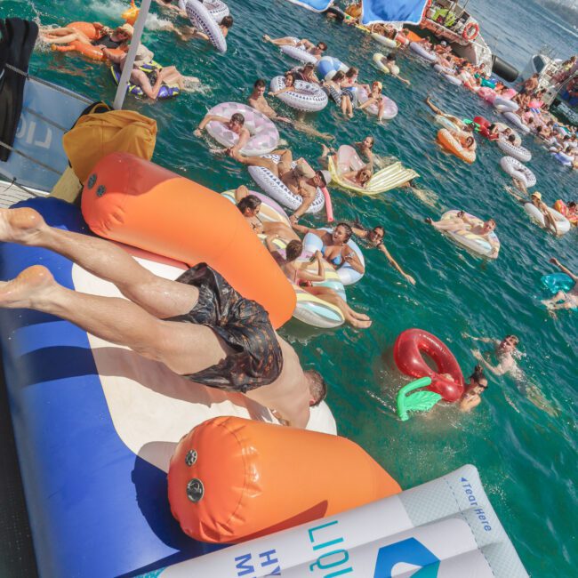 People relax on colorful inflatables in a crowded, sunny pool party. A large "Liquid I.V." canopy and inflatable are visible, with people floating and swimming in bright blue water.