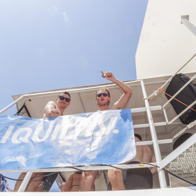 Two people in sunglasses stand on a boat deck behind a blue "LIQUIFY" banner, looking down and smiling at the camera under a clear blue sky. One points upwards, and sunlight brightens the scene.