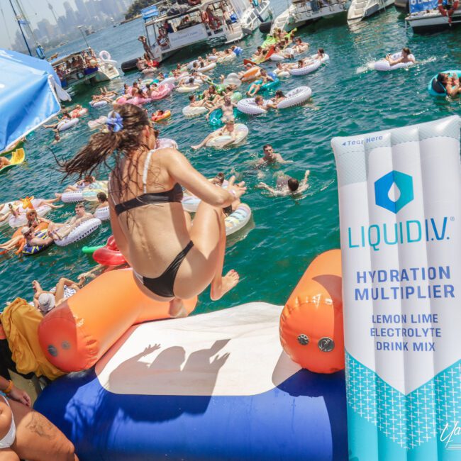 A woman in a bikini jumps off a floating platform into a crowded lake with people on inflatable tubes. A large Liquid I.V. Hydration Multiplier drink mix packet is in the foreground. Boats and a distant city skyline are visible.