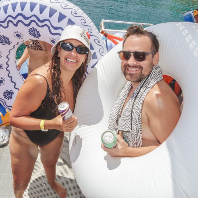 Two people in swimsuits and sunglasses smile at the camera while holding drinks and standing inside inflatable pool rings on a boat, surrounded by water and other people enjoying a sunny day.