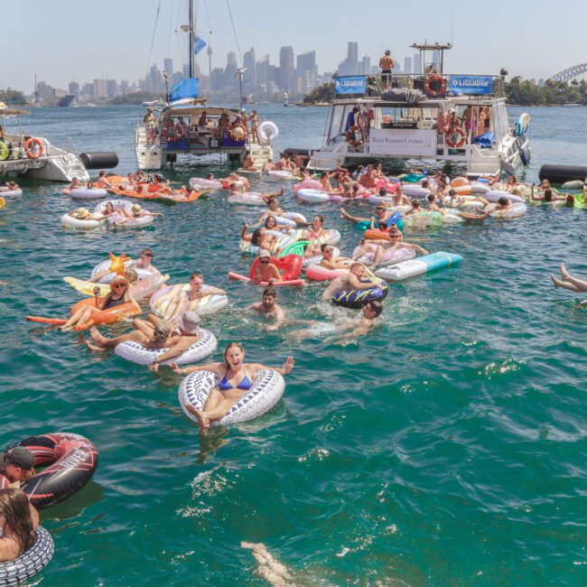 A large group of people relax on colorful inflatable tubes in the water near several boats, with a city skyline and Sydney Harbour Bridge visible in the background, under a sunny sky.