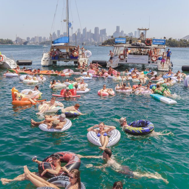 A large group of people relax on inflatables and swim in the water near anchored boats, with a city skyline and the Sydney Harbour Bridge visible in the background on a sunny day.