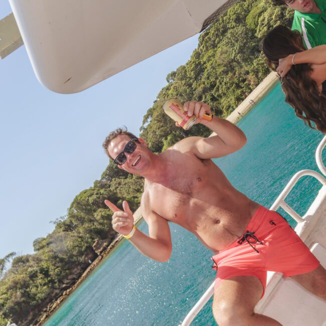 A smiling man in coral swim trunks stands on a boat, holding a hot dog and giving thumbs-up. Two other people stand nearby. The boat is on turquoise water near a forested shoreline under a clear sky.