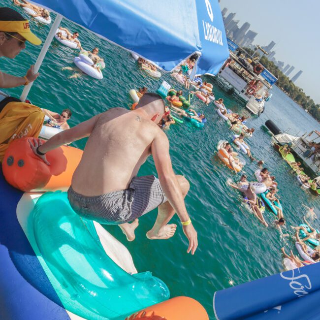 A man in swim trunks slides from a yacht into the water, surrounded by people on pool floats. Blue umbrellas with "Yacht Social" branding provide shade; city buildings are visible in the background.