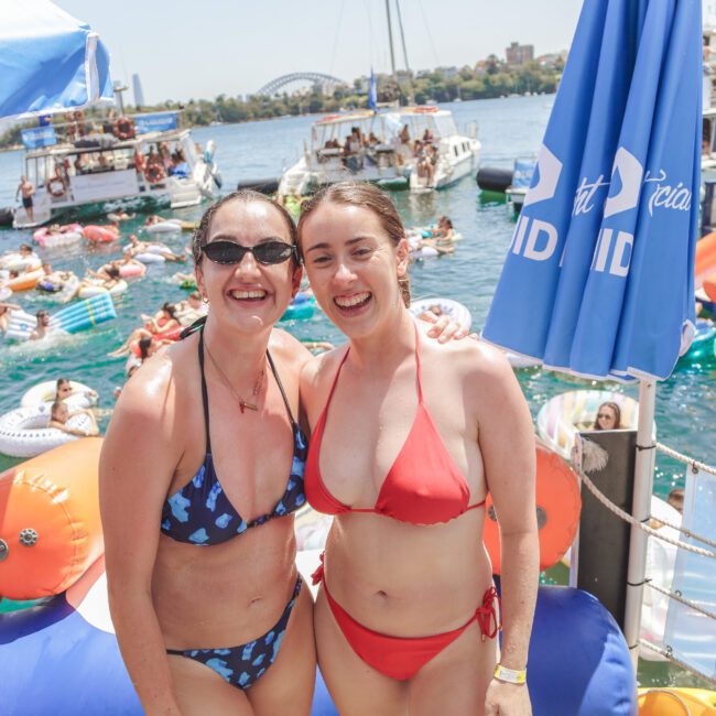 Two women in swimsuits smiling at the camera on a dock by the water, with boats, people, and colorful inflatables in the background on a sunny day.