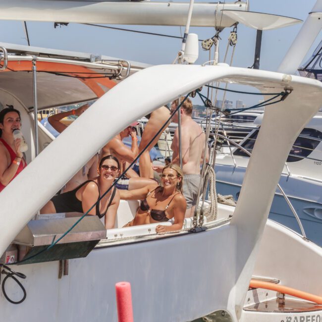 A group of people in swimsuits smiles and relaxes on a sailboat docked at a marina on a sunny day, with other boats and clear blue sky in the background.