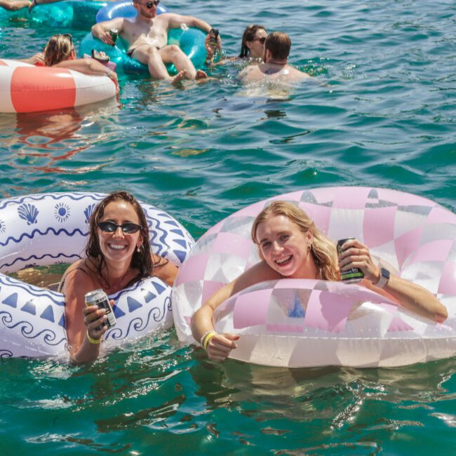 Two women in sunglasses smile while holding drinks and floating in patterned inflatable tubes on a sunny day, surrounded by others relaxing in colorful floats on clear, blue-green water.