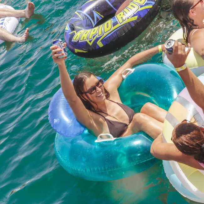 People relax and smile on colorful inflatable tubes, floating in sunny, blue-green water. One woman in sunglasses raises a drink, while others enjoy the water and the summer atmosphere.