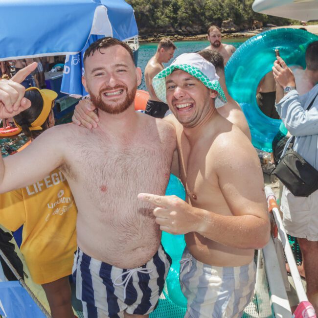 Two smiling men in swim trunks pose together on a boat, surrounded by other people in swimwear, inflatables, and water toys, enjoying a sunny day near the water.