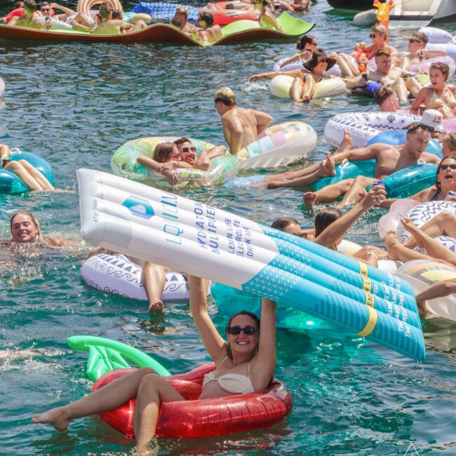 Dozens of people relax and have fun on colorful inflatable pool floats in the water near boats on a sunny day; a woman in sunglasses smiles and holds a large inflatable sunscreen bottle.