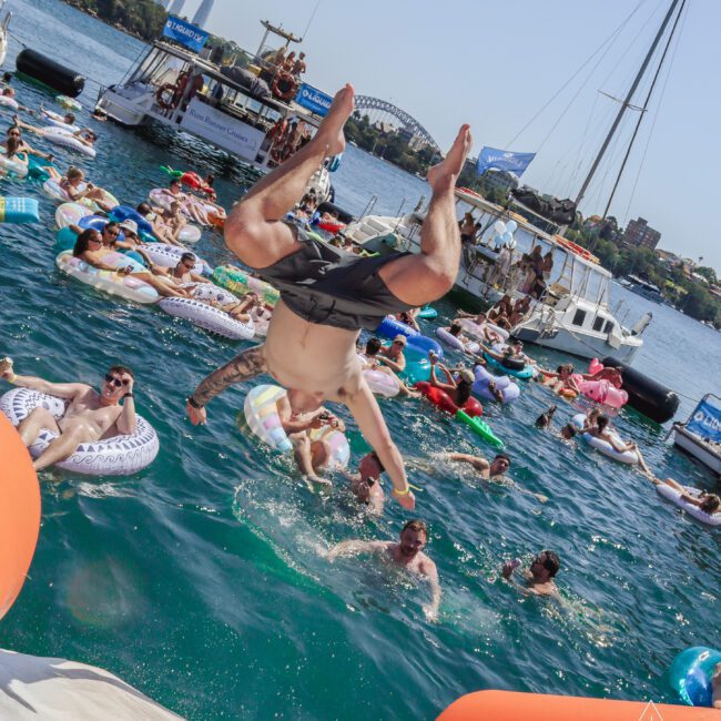 A man does a backflip from an orange waterslide into a bay crowded with people on inflatables; boats and city skyline, including the Sydney Harbour Bridge, are visible in the background on a sunny day.