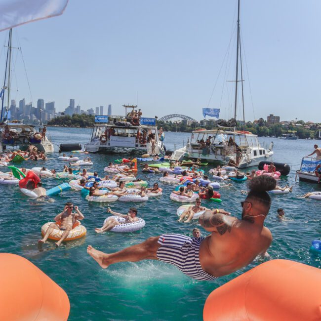 A man in swim trunks slides into the water from an orange float, joining a lively crowd of people on colorful inflatables and boats, with a city skyline and the Sydney Harbour Bridge in the background.