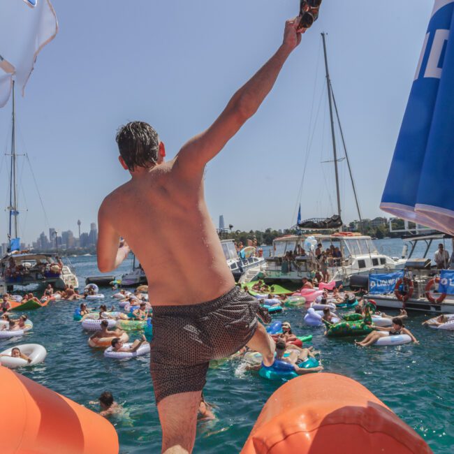 A man in swim trunks leaps off a yacht toward a crowded bay with people on inflatables. Boats and yachts are anchored nearby under a clear, sunny sky. The scene is festive and lively.