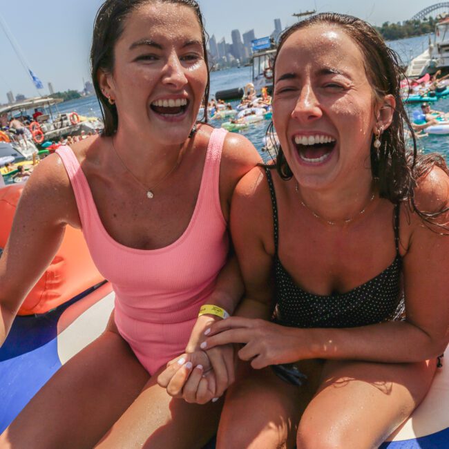 Two young women in swimsuits sit close together on a boat, laughing and holding hands. Behind them, people enjoy the water and city buildings are visible in the distance under a sunny sky.