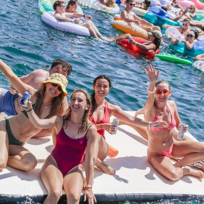 Four people in swimsuits smile and wave while sitting on a floating mat in a sunny, crowded lake. Other people on colorful inflatables relax and enjoy the water in the background.