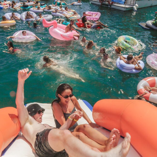 Two people slide into the water from an inflatable on a crowded, sunny lake. Many others float on colorful inflatables near docked boats, enjoying a lively outdoor party atmosphere.