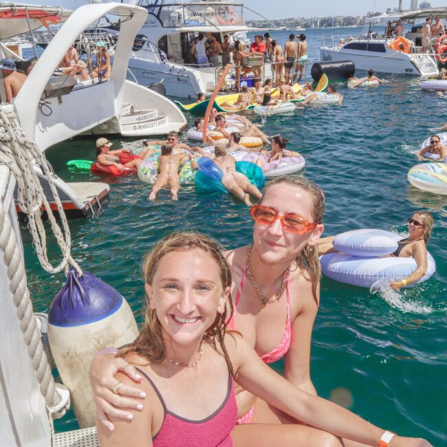 Two women in swimsuits smile at the camera on a boat, with a lively crowd of people swimming, floating on inflatables, and socializing in the water among several boats on a sunny day.