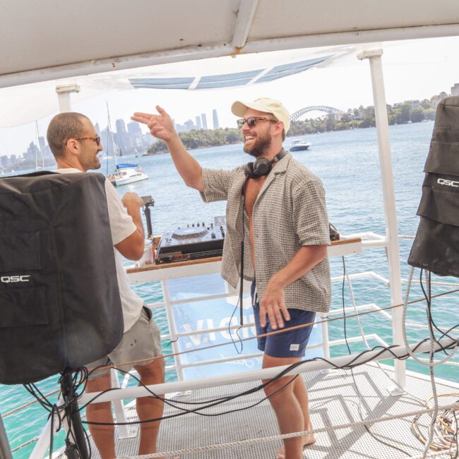 Two men smile and high-five next to DJ equipment on a boat deck, surrounded by large speakers, with a city skyline and a bridge visible across the water in the background.