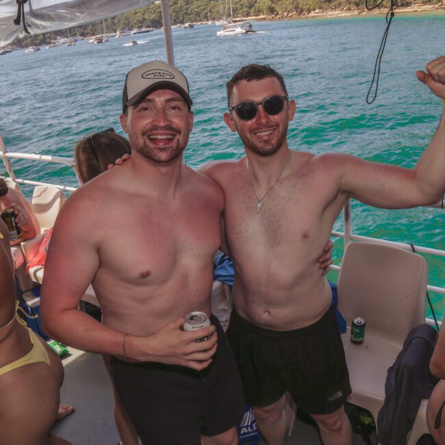 Two smiling men wearing swim trunks and sunglasses pose together on a boat, surrounded by people in swimwear. The boat is on clear blue water with trees and other boats visible in the background.