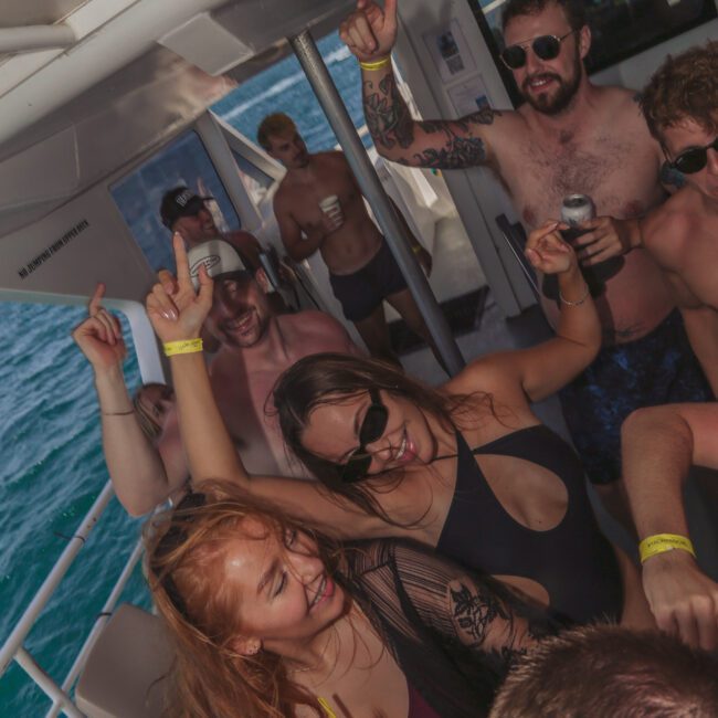 A group of young adults in swimsuits dance, laugh, and enjoy drinks on a boat, with the ocean visible in the background. Some wear sunglasses and wristbands, creating a lively, festive atmosphere.
