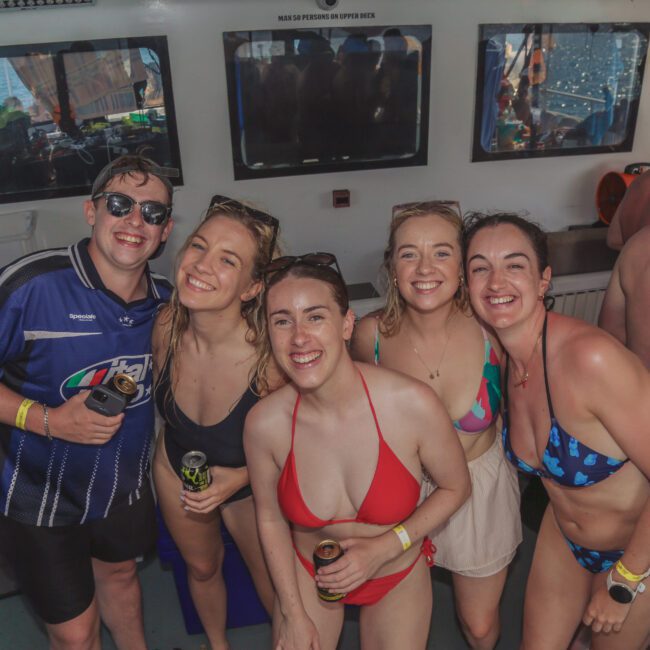 A group of five young adults in swimwear smile and pose for a photo on a crowded party boat, holding drinks. Other people are mingling in the background, and the sea is visible through the windows.