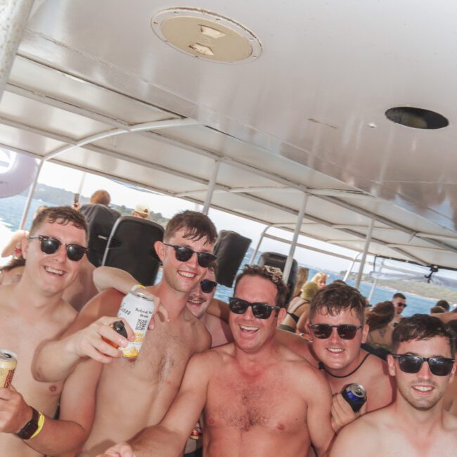A group of six smiling men, most shirtless and wearing sunglasses, pose with drinks in hand on a boat. Other people are seen in the background under a shaded area. The atmosphere is festive and relaxed.