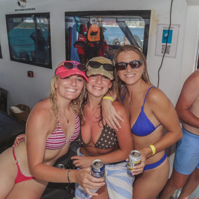 Three women in swimsuits and sunglasses smile and pose together on a boat, each holding a drink. Other people are in the background, with one man in swim trunks and sunglasses smiling at the camera. It's a sunny day on the water.