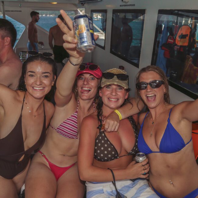 A group of smiling young adults in swimsuits celebrate on a boat, holding drinks and posing for the camera with arms raised. Others in swimwear enjoy the party in the background near windows and the water.