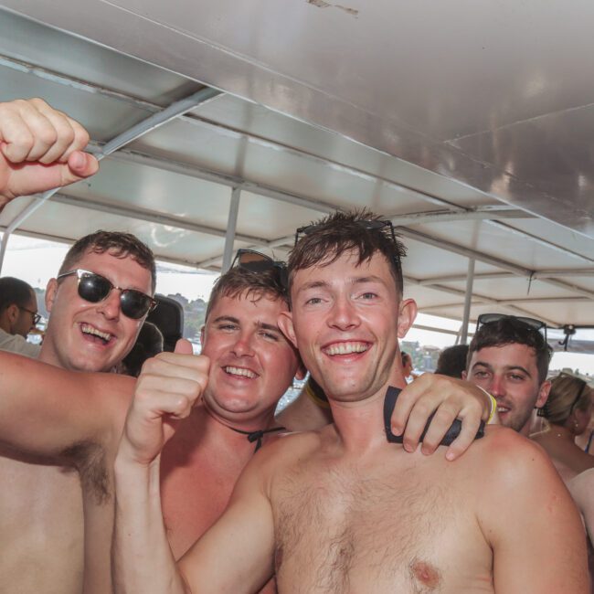 A group of young adults in swimsuits smiling and raising their fists in celebration on a boat, with more people and water visible in the background. The atmosphere appears lively and festive.