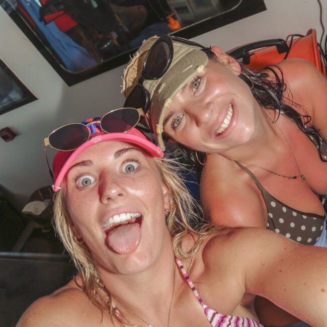 Two women in swimsuits and caps take a playful selfie on a boat, smiling and making faces. Sunglasses rest on their caps, and boating equipment is visible in the background. Logo reads "Yacht Social.