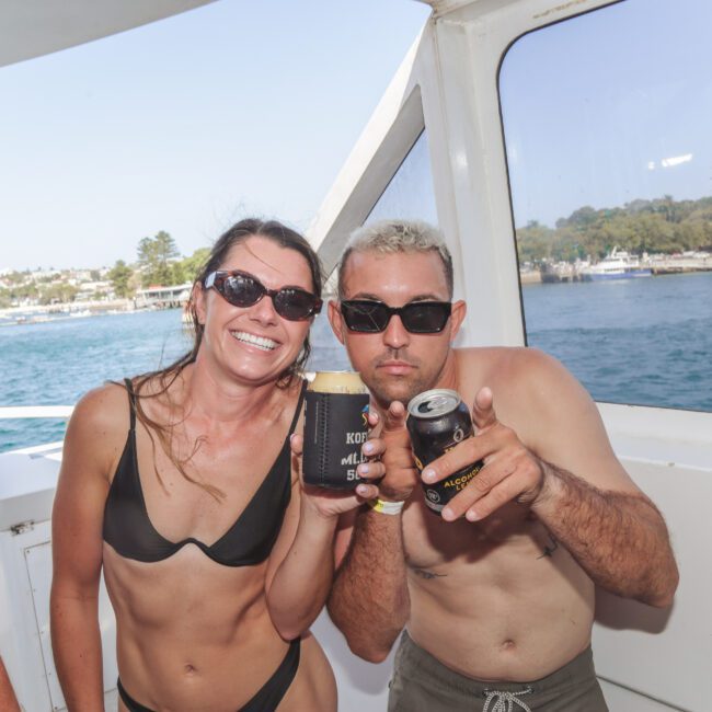 A smiling woman in a black bikini and a man in sunglasses pose together on a boat, holding drinks. The sea and coastline with trees and houses are visible in the background under a clear sky.