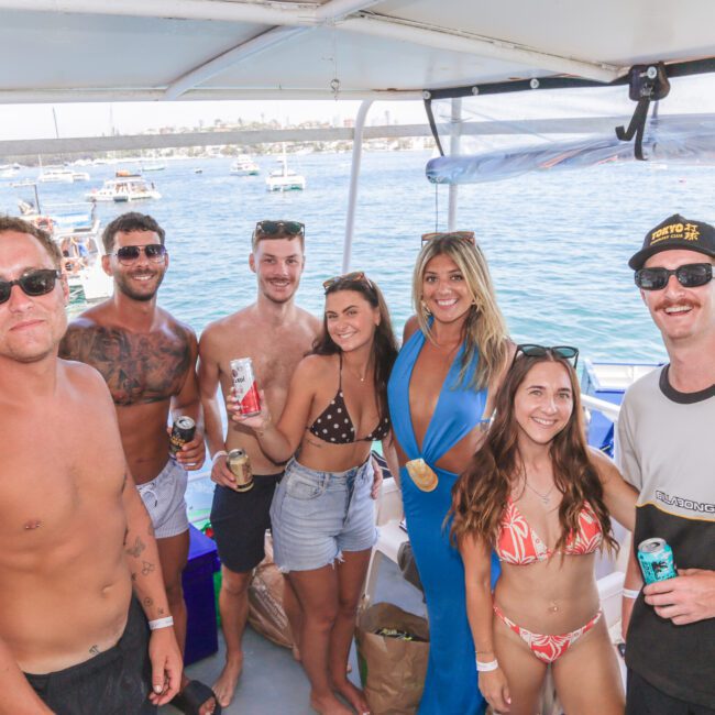 A group of seven smiling young adults in swimwear and summer attire stand together on a boat deck, holding drinks, with a marina and other boats visible on the blue water in the background.